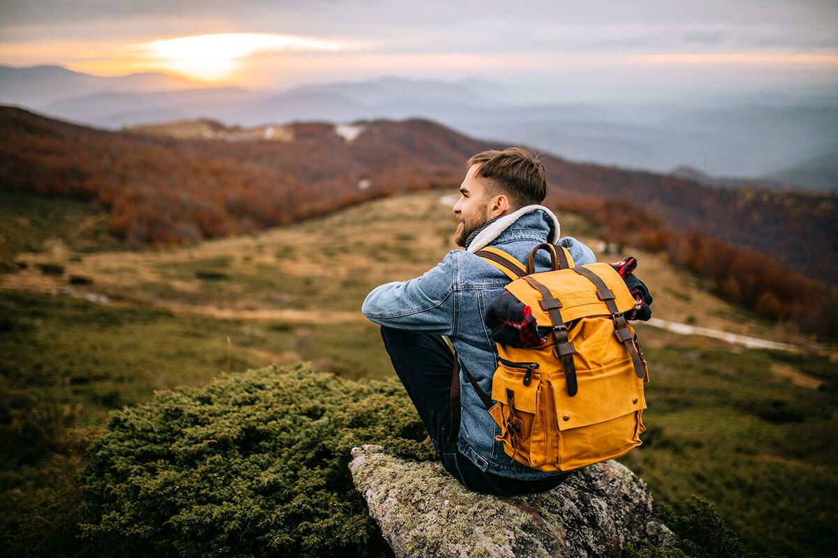 A man wearing a backpack sits on a rock, enjoying a scenic view of rolling hills and a sunset.