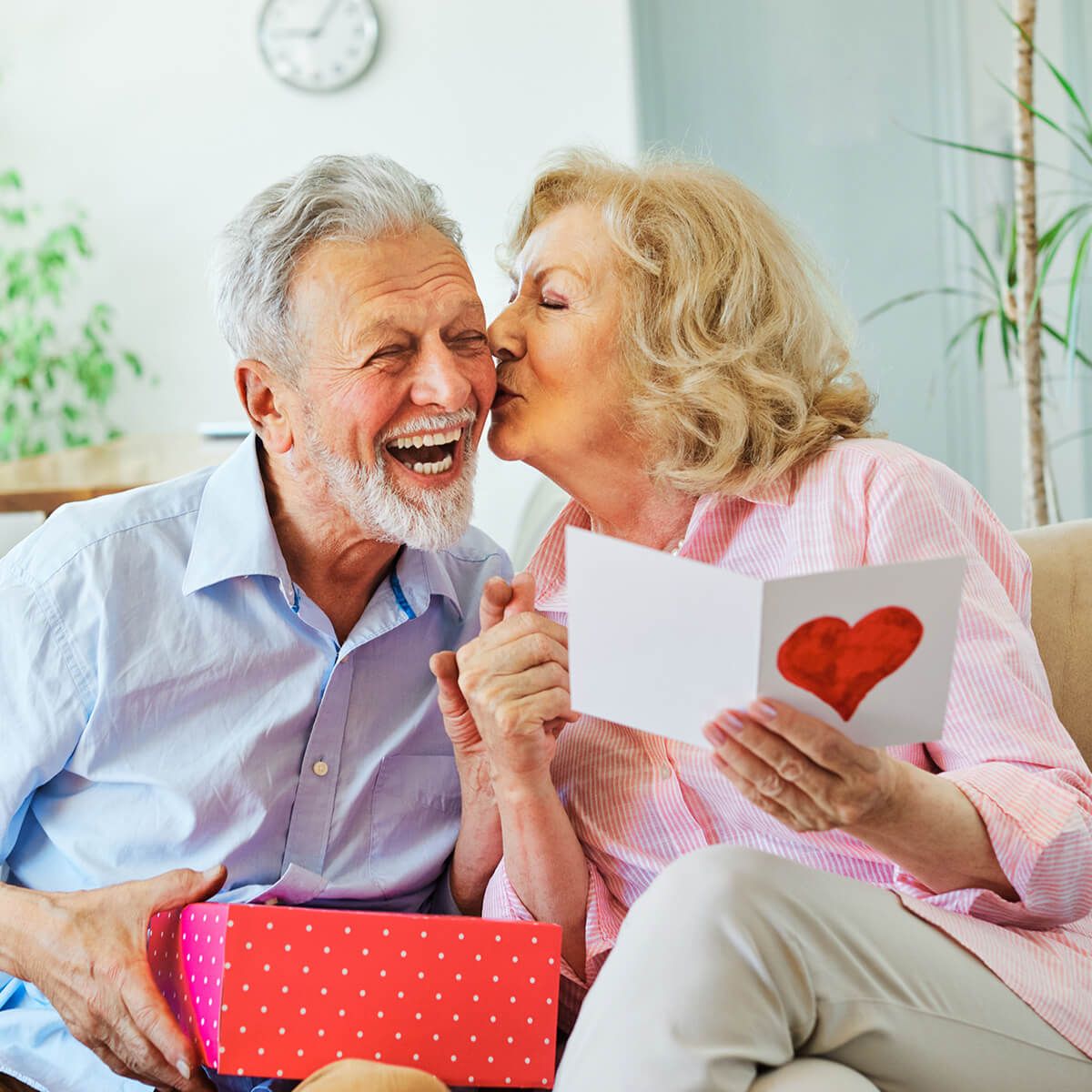 An elderly couple sitting on a couch, exchanging smiles as the woman kisses the man on the cheek while holding a heartfelt card and a gift box.