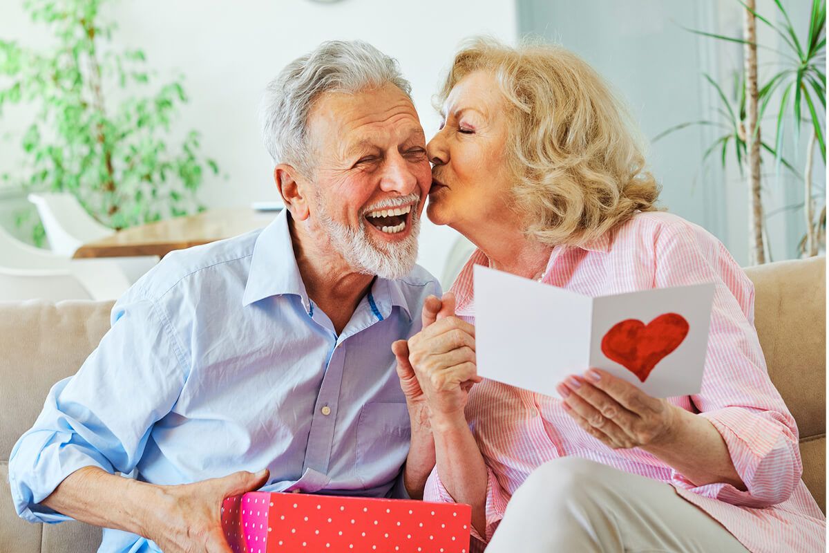 An elderly couple sitting on a couch, exchanging smiles as the woman kisses the man on the cheek while holding a heartfelt card and a gift box.