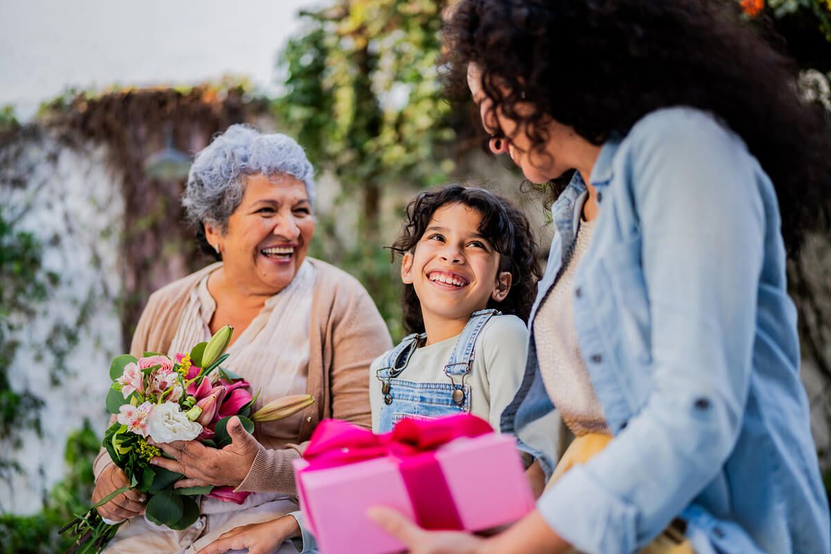 Three generations celebrating Mother’s Day with joy and gifts Grandmother, mother, and daughter smiling and laughing together while holding flowers and a pink gift box