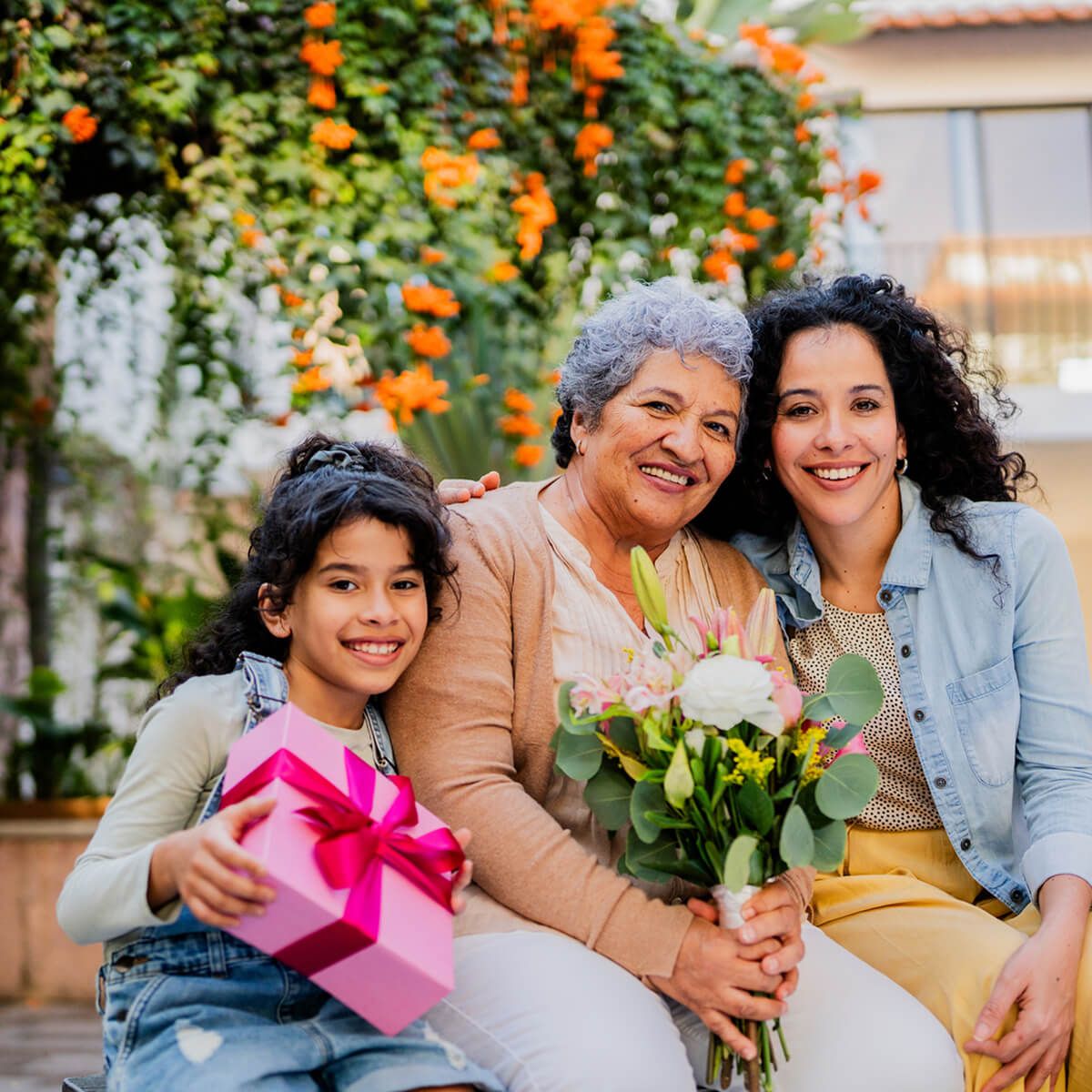 Smiling girl, mother, and grandmother sitting together outdoors with flowers and a pink gift box